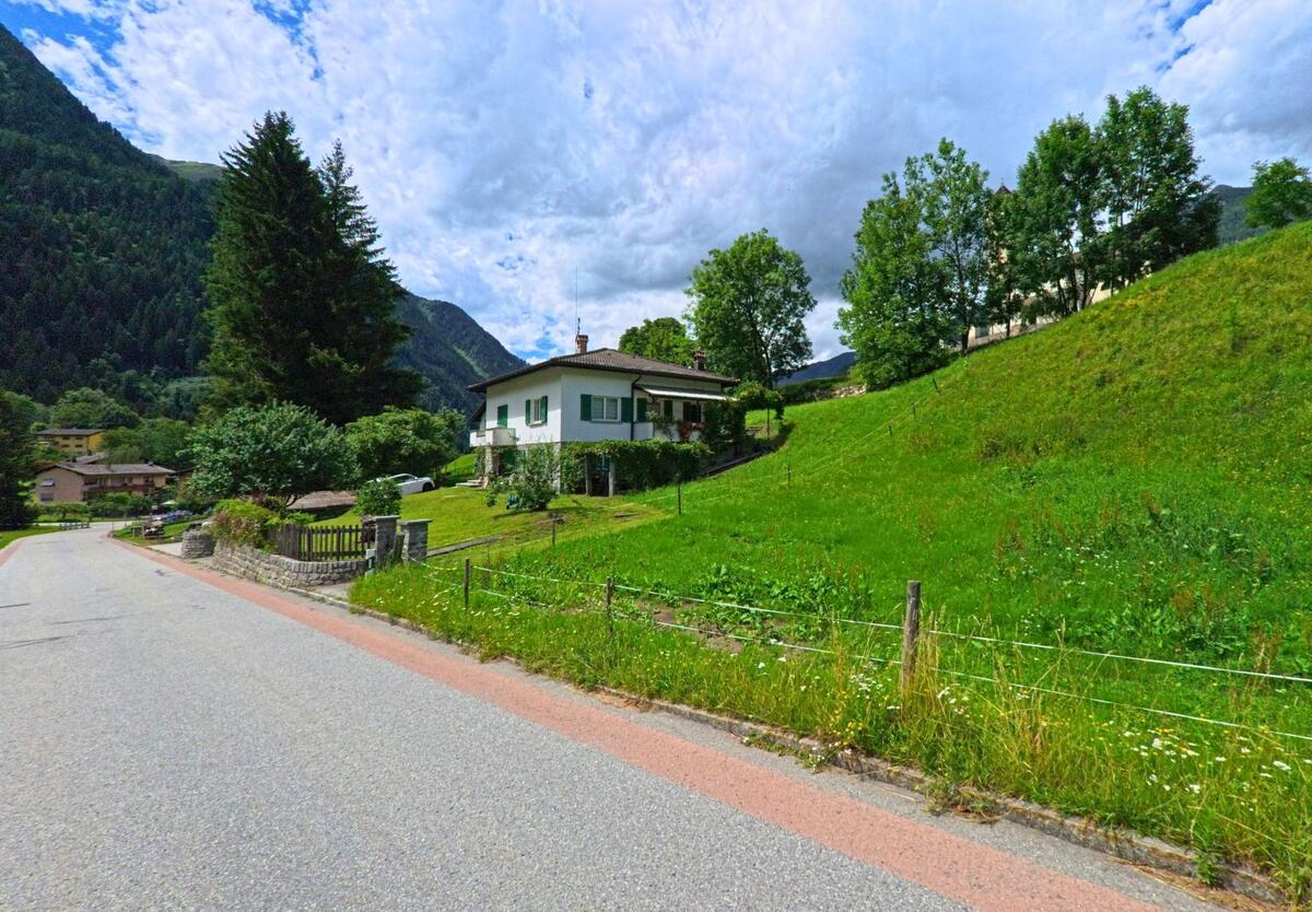 Ein Einfamilienhaus mit Balkon und grüner Umgebung steht am Hang in Prato (Leventina) (6773, Kanton Tessin), eingerahmt von Wiesen, Bäumen und Berglandschaft.