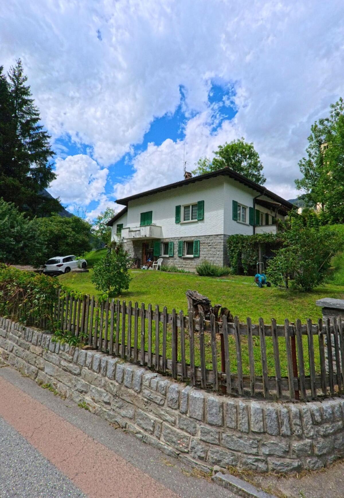 Ein zweistöckiges Haus mit grünen Fensterläden und kleinem Garten steht in Prato (Leventina) (6773, Kanton Tessin) an einer ruhigen Strasse mit Steinmauer und Holzzaun.
