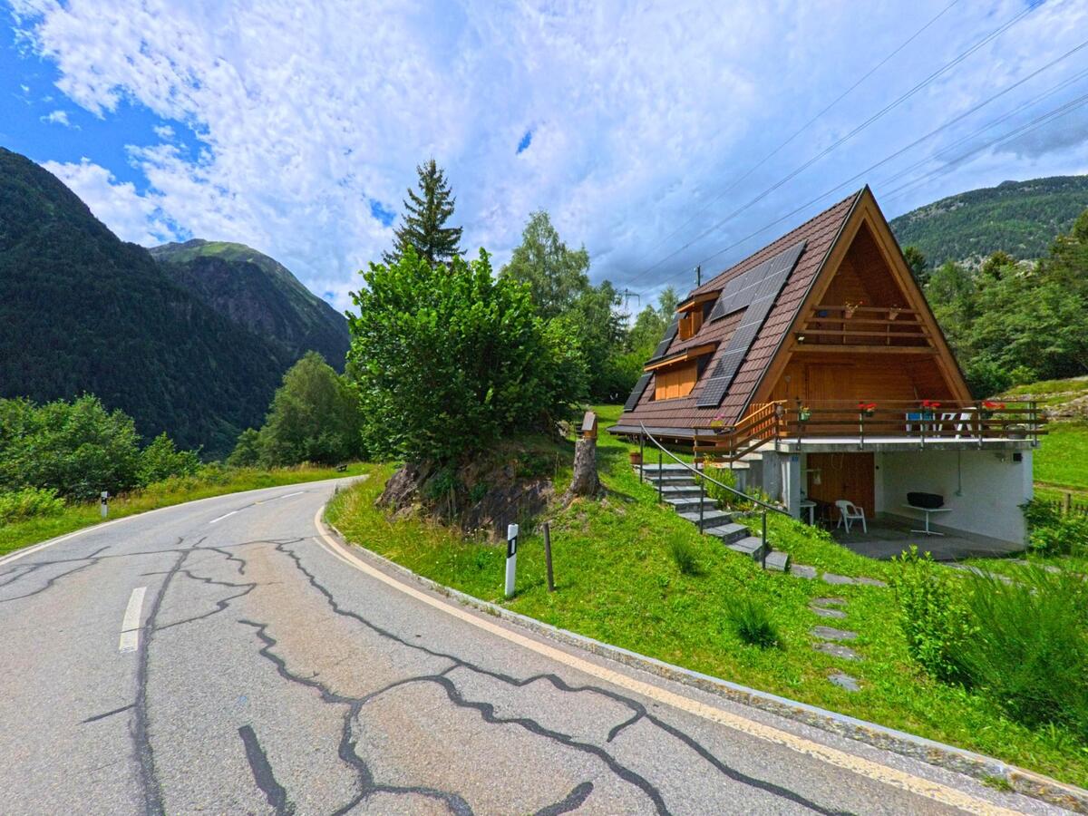 Ein Holzhaus mit steilem Dach und Solarpanelen steht an einer kurvigen Strasse in Prato (Leventina) (6773, Kanton Tessin), umgeben von grüner Landschaft und bewaldeten Berghängen.