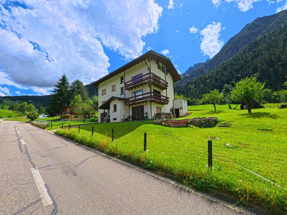 Ein mehrstöckiges Haus mit Holzbalkonen steht am Rand einer Landstrasse in Prato (Leventina) (6773, Kanton Tessin), umgeben von grüner Wiese und bewaldeten Bergen.