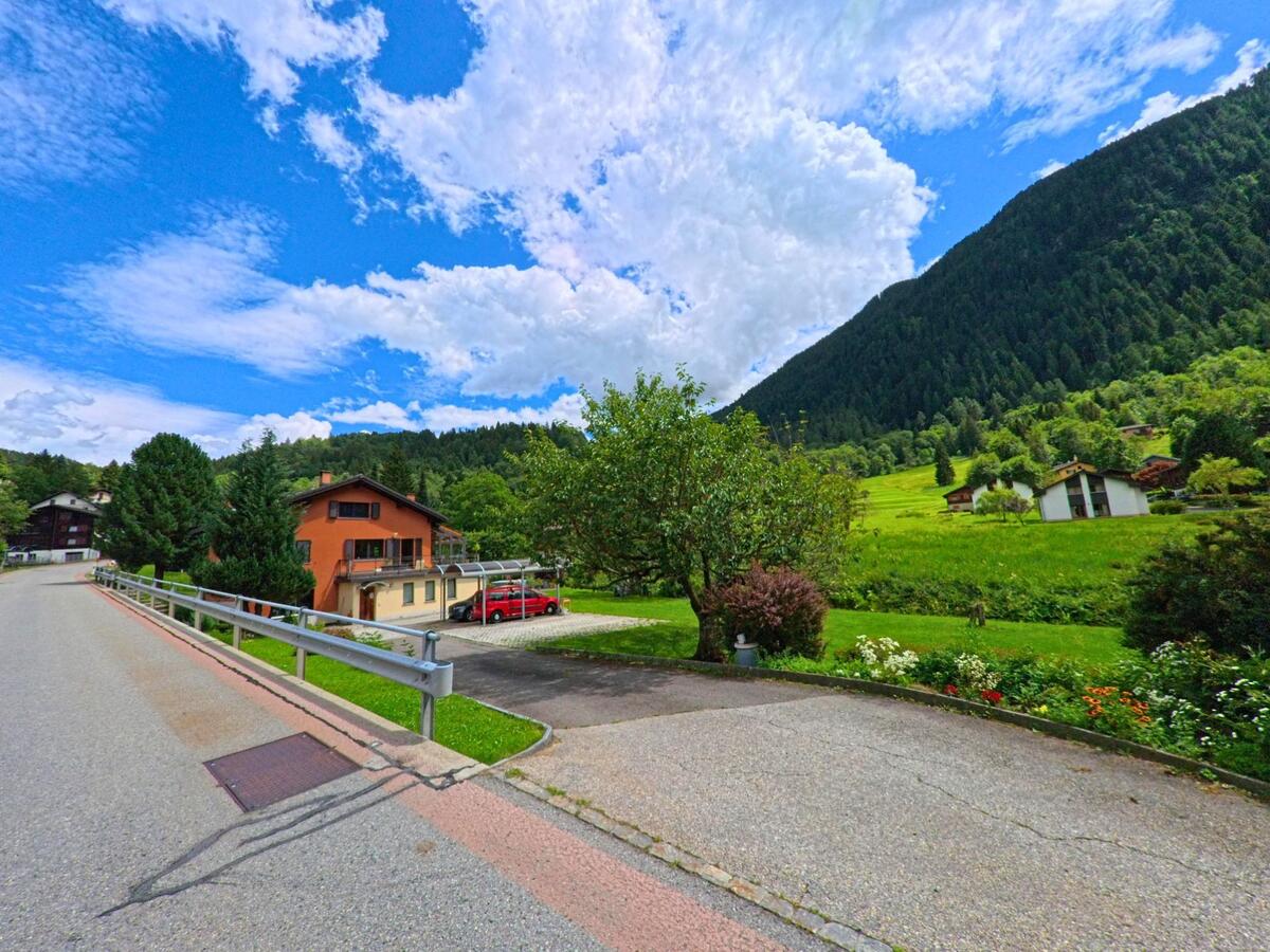 Ein orangefarbenes Haus mit Carport steht an einer ruhigen Strasse in Prato (Leventina) (6773, Kanton Tessin), umgeben von Wiesen, Bäumen und einer hügeligen Berglandschaft.