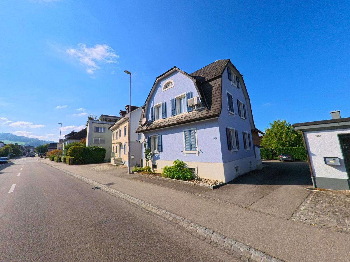 Ein hellblaues Wohnhaus mit geschlossenen Fensterläden steht in Pratteln (4133, Kanton Basel-Landschaft) entlang einer langen Strasse, die sich ruhig durch das Wohnquartier zieht.