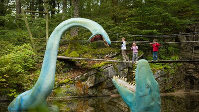 Im Préhisto-Parc et Grottes de Réclère in Réclère (2912, Kanton Waadt) überqueren Kinder eine Hängebrücke über einem Teich mit grossen Dinosaurierfiguren inmitten eines bewaldeten Geländes.Im Préhisto-Parc et Grottes de Réclère in Réclère (2912, Kanton Waadt) überqueren Kinder eine Hängebrücke über einem Teich mit grossen Dinosaurierfiguren inmitten eines bewaldeten Geländes.
