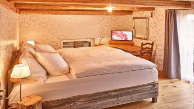 In einem stilvoll eingerichteten Zimmer des Premium BnB Buntekatze in Russikon steht ein grosses Bett mit weisser Bettwäsche und rustikalem Holzrahmen, daneben ein kleiner Schreibtisch mit Fernseher und Stuhl unter freigelegten Deckenbalken.