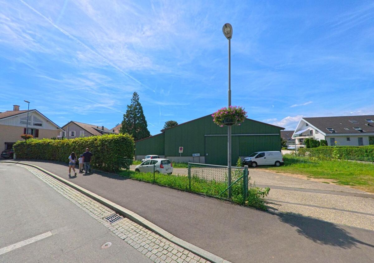 Zwei Personen spazieren auf einem Gehweg in Préverenges (1028, Kanton Waadt) vorbei an einem gruenen Lagergebaeude und angrenzenden Wohnhaeusern.