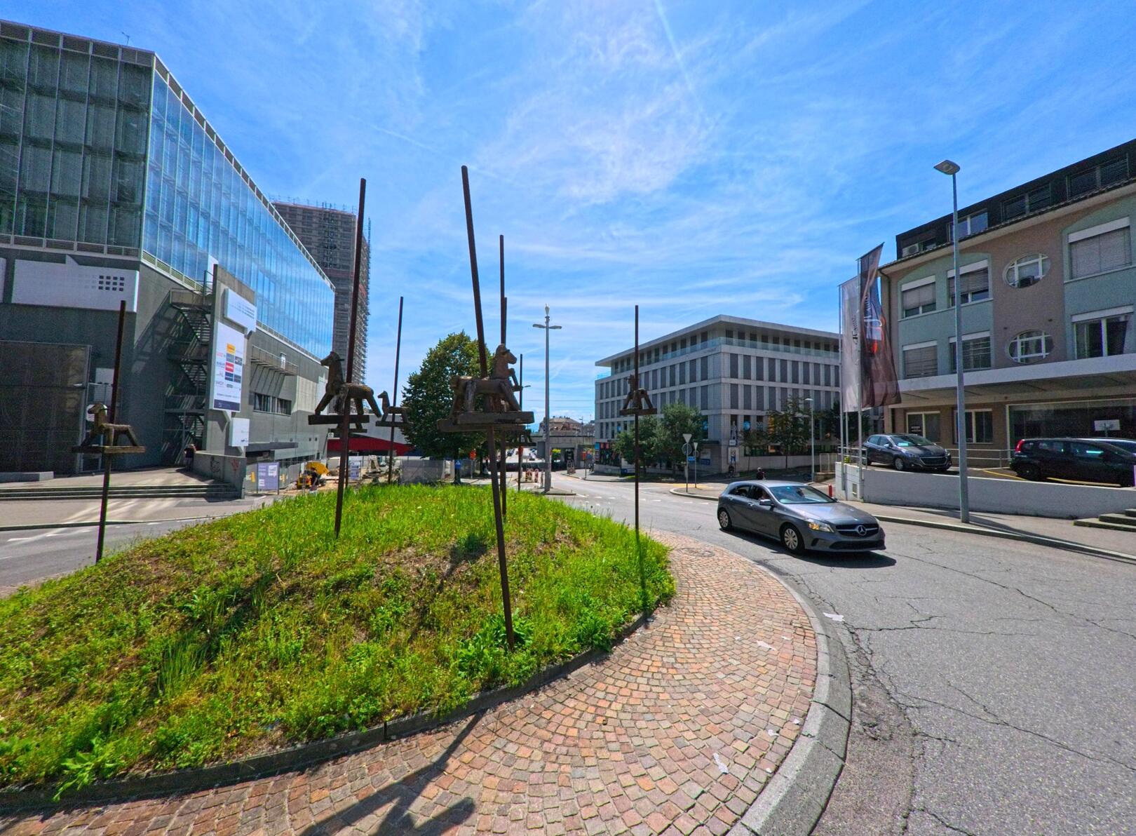 Auf einem Kreisverkehr in Prilly (1008, Kanton Waadt) stehen mehrere Metallskulpturen in Form von Reitpferden, umgeben von modernen Gebaeuden und einer befahrenen Strasse.