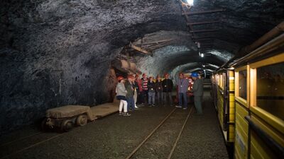 In einem Stollen des Pro Gonzenbergwerks in Sargans (7320, Kanton St. Gallen) steht eine Gruppe Besucher mit Helmen neben einer gelben Grubenbahn und hört einer Führung zu.
