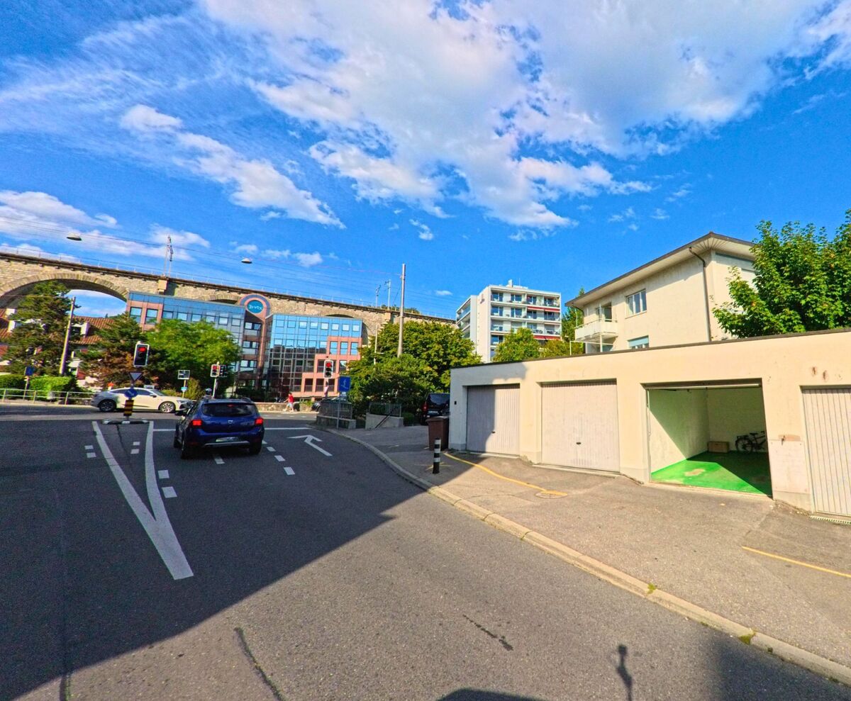 Ein Strassenzug in Pully (1009, Kanton Waadt) zeigt Garagen, Wohnhaeuser und ein modernes Buerohaus, im Hintergrund verlaeuft ein steinernes Eisenbahnviadukt.