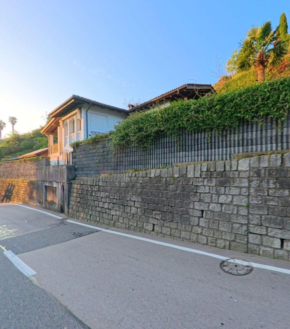 Entlang einer schmalen Strasse in Pura (6987, Kanton Tessin) verläuft eine hohe Natursteinmauer vor Wohnhäusern mit Gärten am sonnigen Hang.