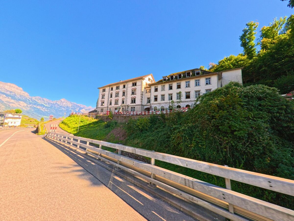 Ueber einer ansteigenden Strasse in Quarten (8877, Kanton St. Gallen) steht ein grosses, helles Gebaeude mit Bergblick und dichter Vegetation im Vordergrund.