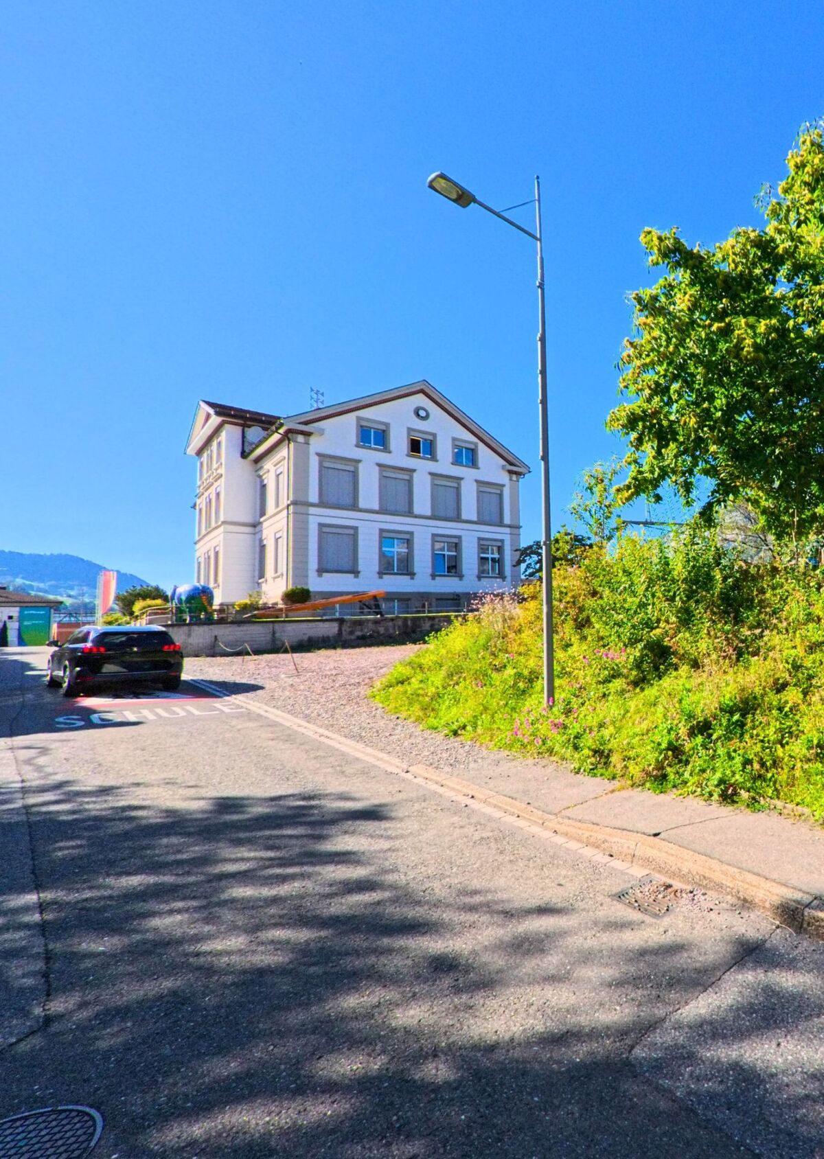 Ein helles Schulgebaeude in Quarten (8877, Kanton St. Gallen) steht an einer ruhigen Strasse mit deutlicher Markierung und Blick auf die umliegenden Huegel.