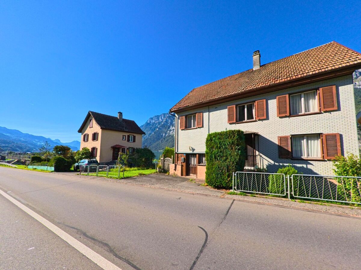 Zwei Wohnhaeuser mit braunen Fensterlaeden stehen in Quarten (8882, Kanton St. Gallen) entlang einer sonnigen Strasse mit Blick auf die Berge.