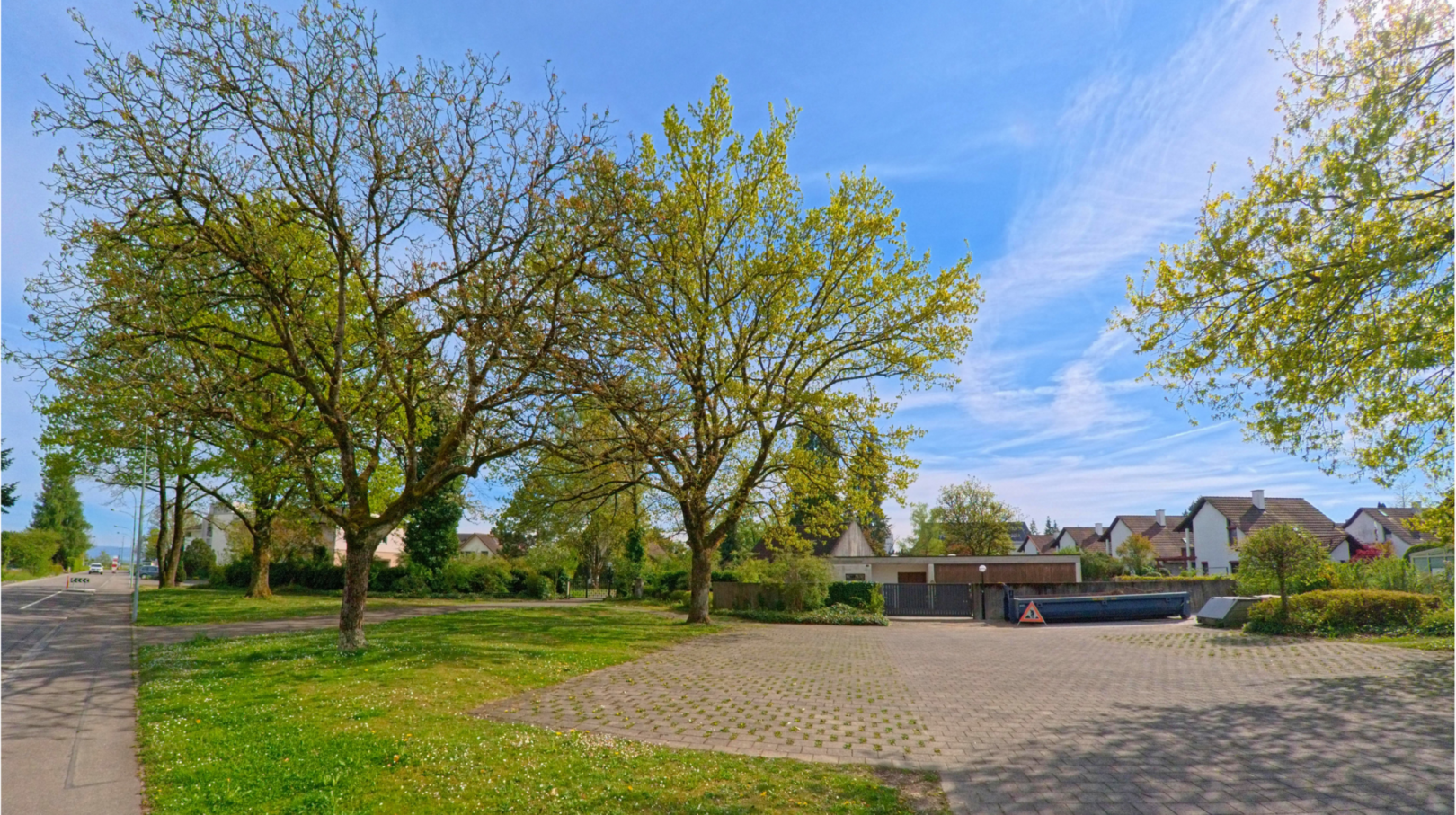 In Rafz im Kanton Zürich mit der PLZ 8197 liegt ein gepflegter Grünbereich mit grossen Bäumen und einem angrenzenden Platz vor einer Wohnsiedlung.