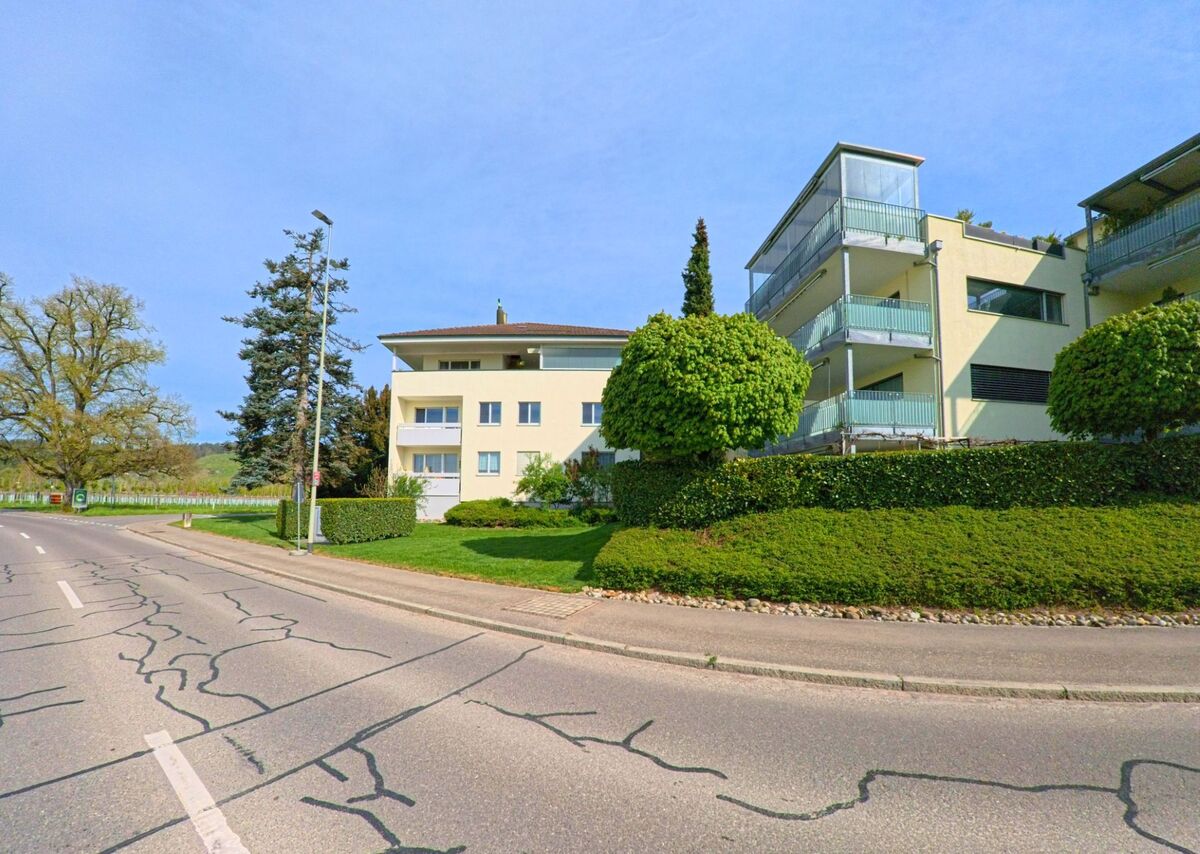 In Rafz im Kanton Zürich, PLZ 8197, stehen moderne Wohngebäude mit Balkonen an einer leicht ansteigenden Strasse, umgeben von gepflegten Grünflächen.
