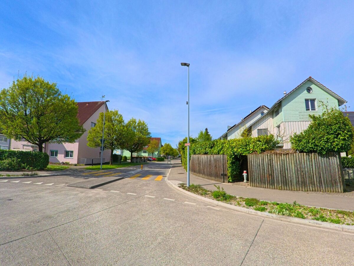  In Rafz im Kanton Zürich mit der PLZ 8197 verläuft eine ruhige Wohnstrasse mit bunt gestrichenen Häusern, Vorgärten und einem Fussgängerstreifen.