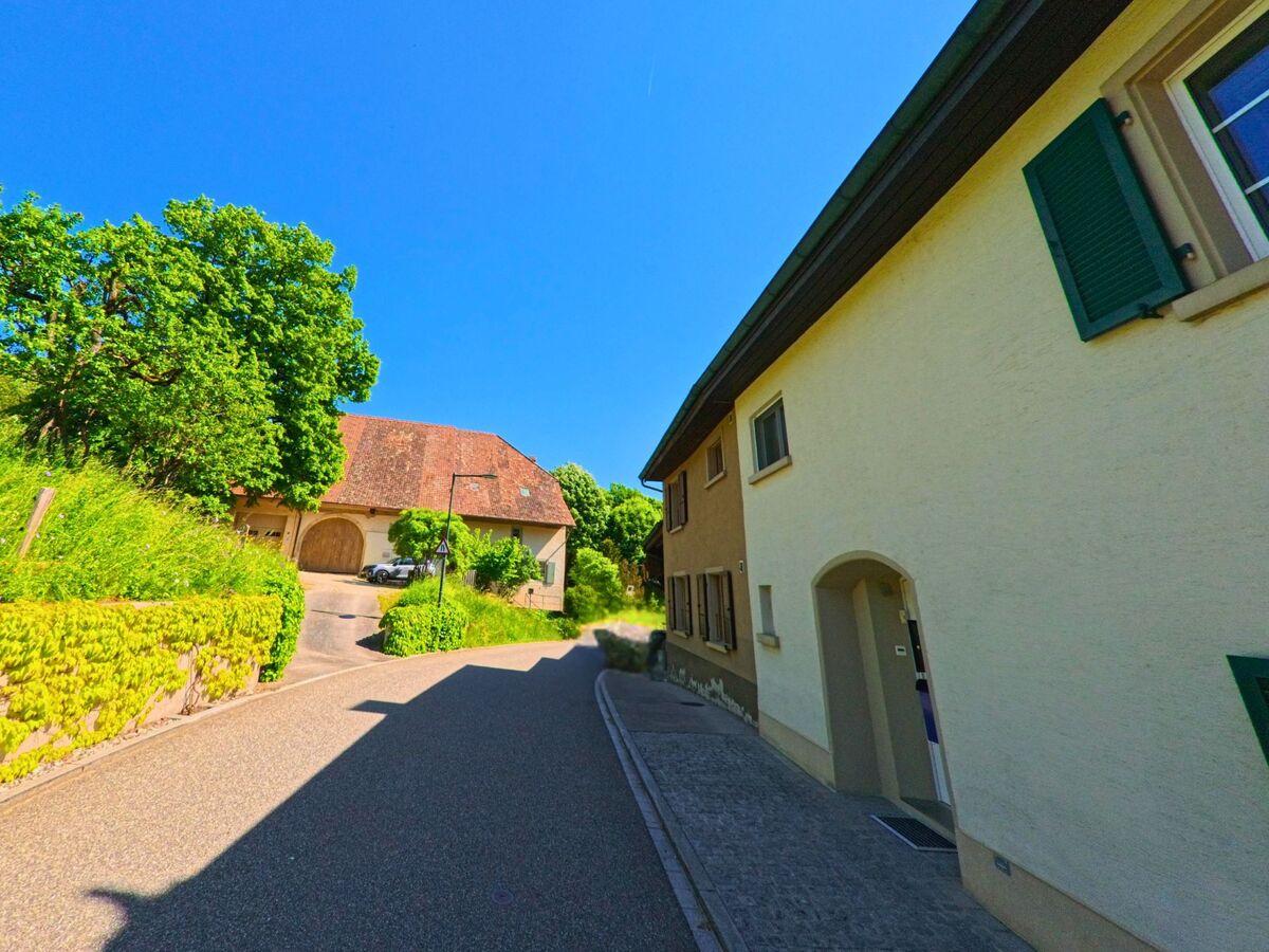 In Ramlinsburg (4433, Kanton Baselland) verläuft eine schmale Strasse zwischen gepflegten Häusern und einer Scheune mit grossen Holztoren in ländlicher Umgebung.