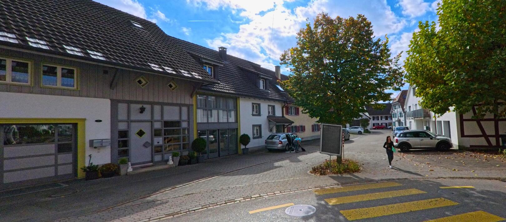 In einer kleinen Platzsituation von Ramsen (8262, Kanton Schaffhausen) stehen unter herbstlichen Baumen mehrere Wohnhaeuser, waehrend Menschen zwischen geparkten Autos entlanggehen.