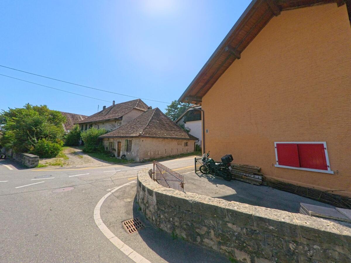 An einer Strassenkurve in Rances (1439, Kanton Waadt) stehen alte Bauernhaeuser mit verwitterten Fassaden neben einem neueren orangefarbenen Gebaeude.