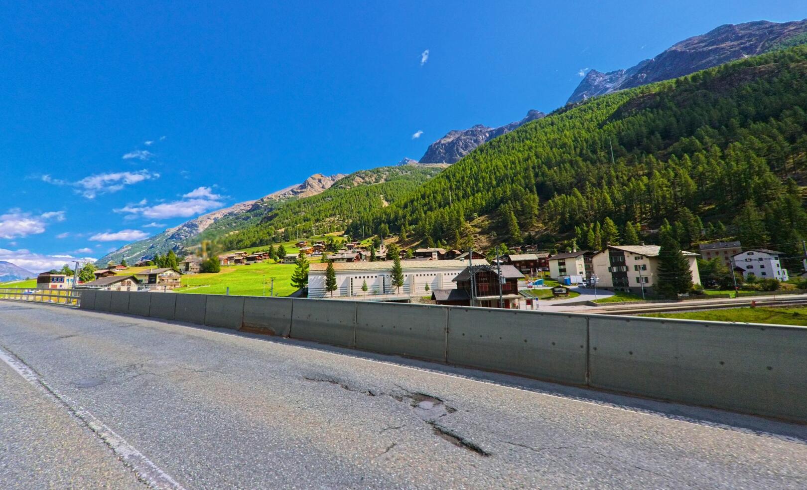 Von der Strasse aus bietet sich ein Blick auf das Dorf Randa (3928, Kanton Wallis), das sich am Hang unterhalb dichter Waelder und felsiger Gipfel ausbreitet.