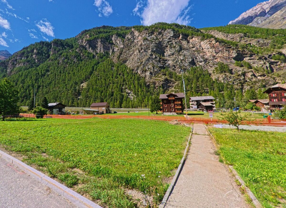 Ein schmaler Fussweg fuehrt in Randa (3928, Kanton Wallis) ueber eine Wiese zu Wohnhaeusern, die am Fusse einer steilen, bewaldeten Felswand liegen.