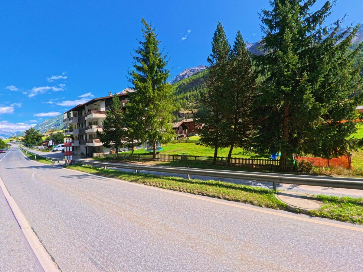 An der Hauptstrasse von Randa (3928, Kanton Wallis) stehen Mehrfamilienhaeuser und Tannenbaeume vor der Kulisse der bewaldeten Berghaenge.