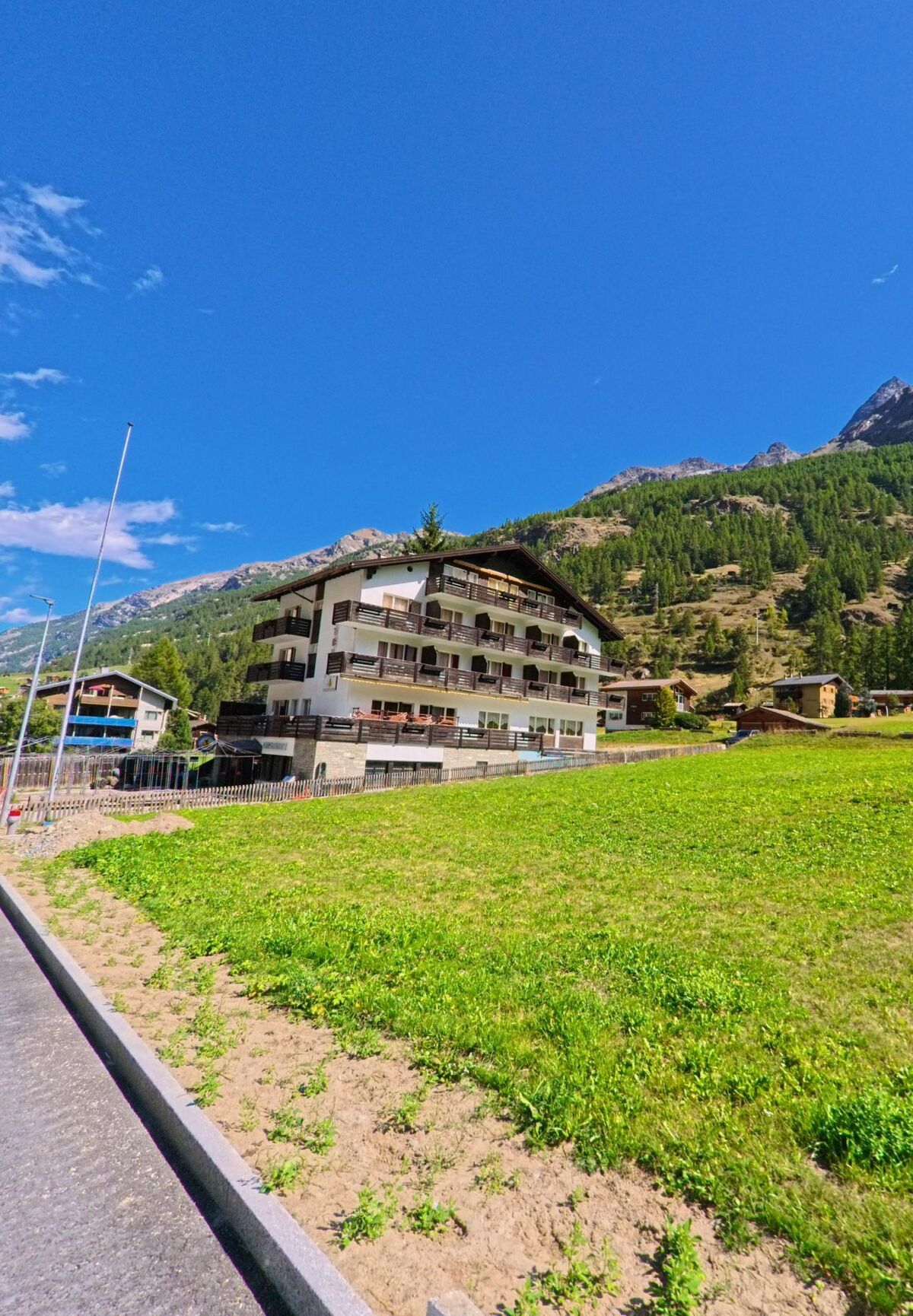 Ein mehrstöckiges Wohnhaus mit Balkonen steht in Randa (3928, Kanton Wallis) vor dem bewaldeten Berghang unter blauem Himmel.