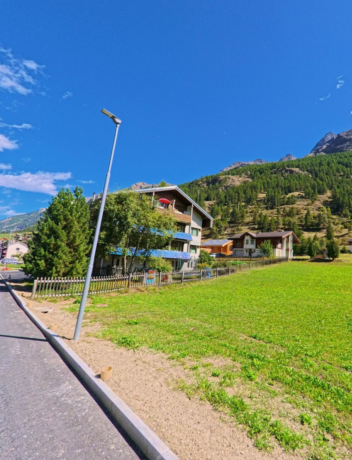 In Randa (3928, Kanton Wallis) stehen moderne Wohnhaeuser mit Gaerten am Hang, umgeben von Wiesen und bewaldeten Bergen.