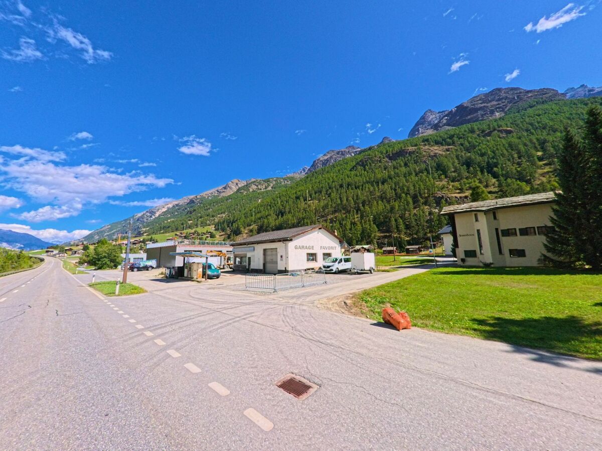 Am Ortseingang von Randa (3928, Kanton Wallis) liegen eine Werkstatt und Wohnhaeuser neben der Hauptstrasse, eingerahmt von bewaldeten Berghaengen.