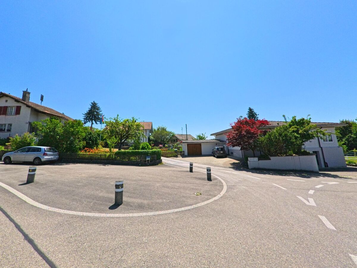 In Rapperswil (3053, Kanton Bern) führt eine ruhige Straße zu einem Kreisverkehr, umgeben von Häusern und gepflegten Gärten, während ein Auto den Kreisverkehr passiert.