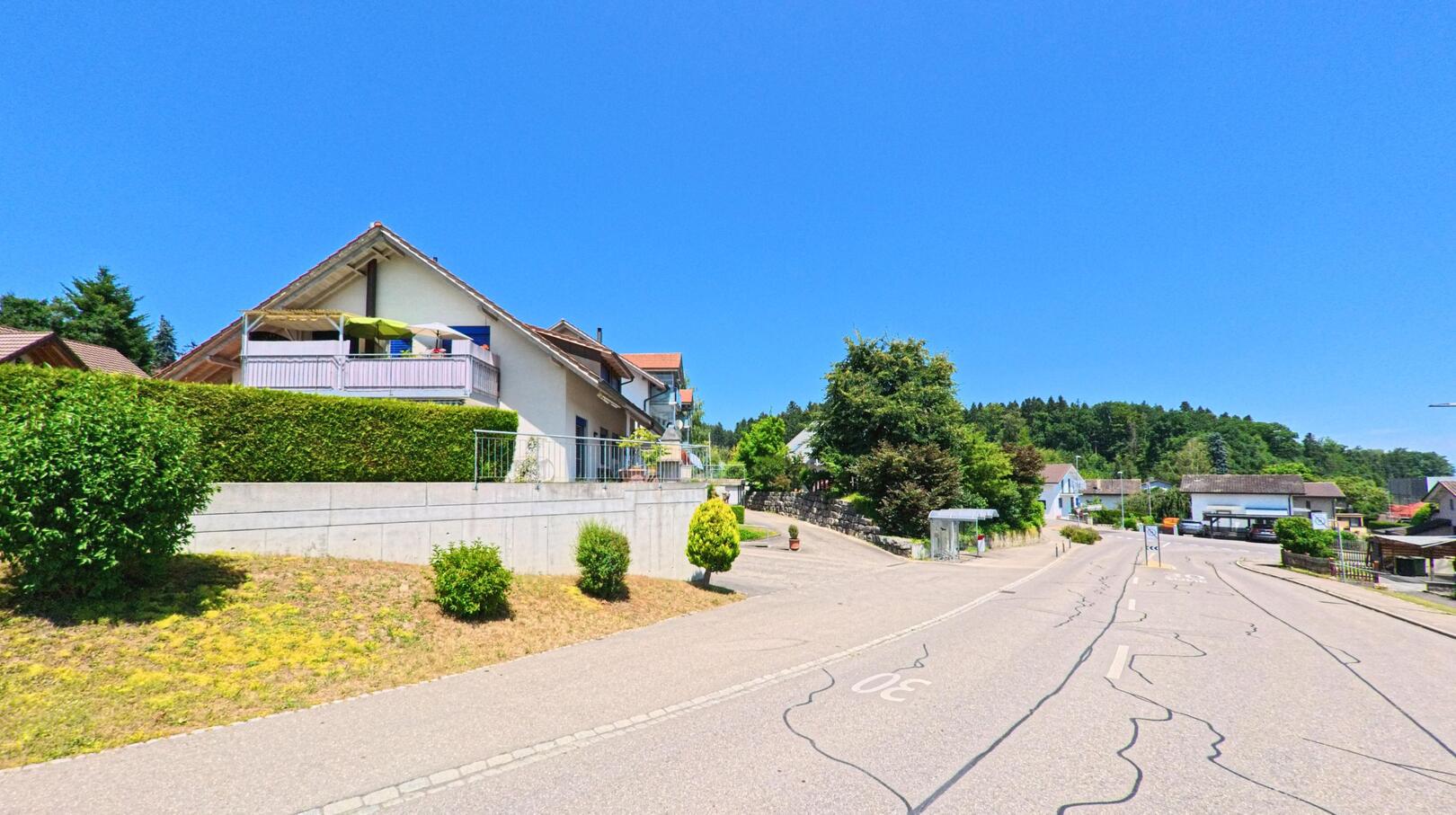 In Rapperswil (3053, Kanton Bern) führt eine ruhige Strasse an gepflegten Häusern vorbei, mit einem klaren Blick auf den Wald im Hintergrund und einer Geschwindigkeitsbegrenzung von 30 km/h.