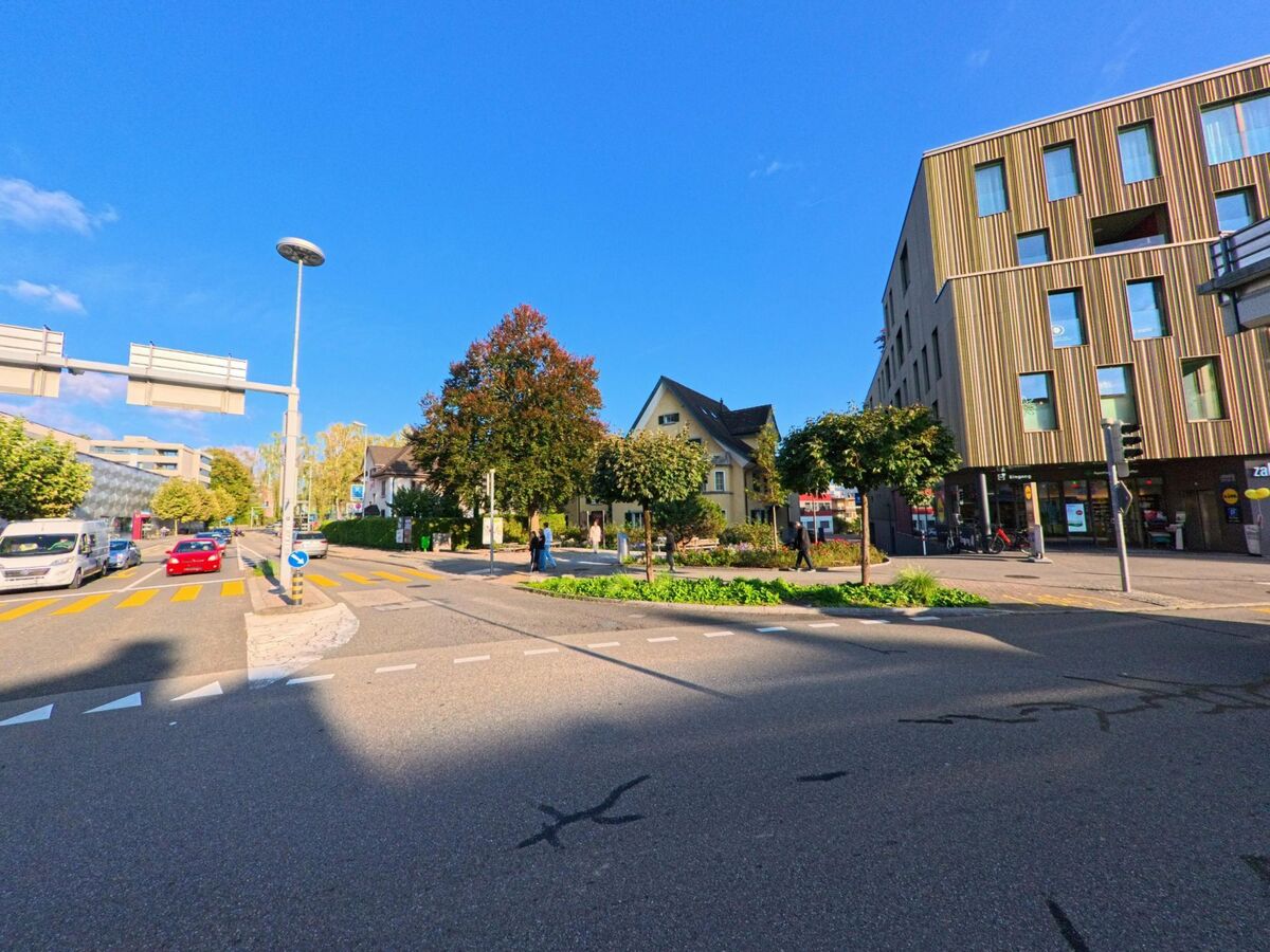 In Rapperswil-Jona (8640, Kanton St. Gallen) liegt eine Kreuzung mit klar markierten Fussgaengerstreifen zwischen modernen und traditionellen Gebaeuden sowie kleinen Baumanpflanzungen.