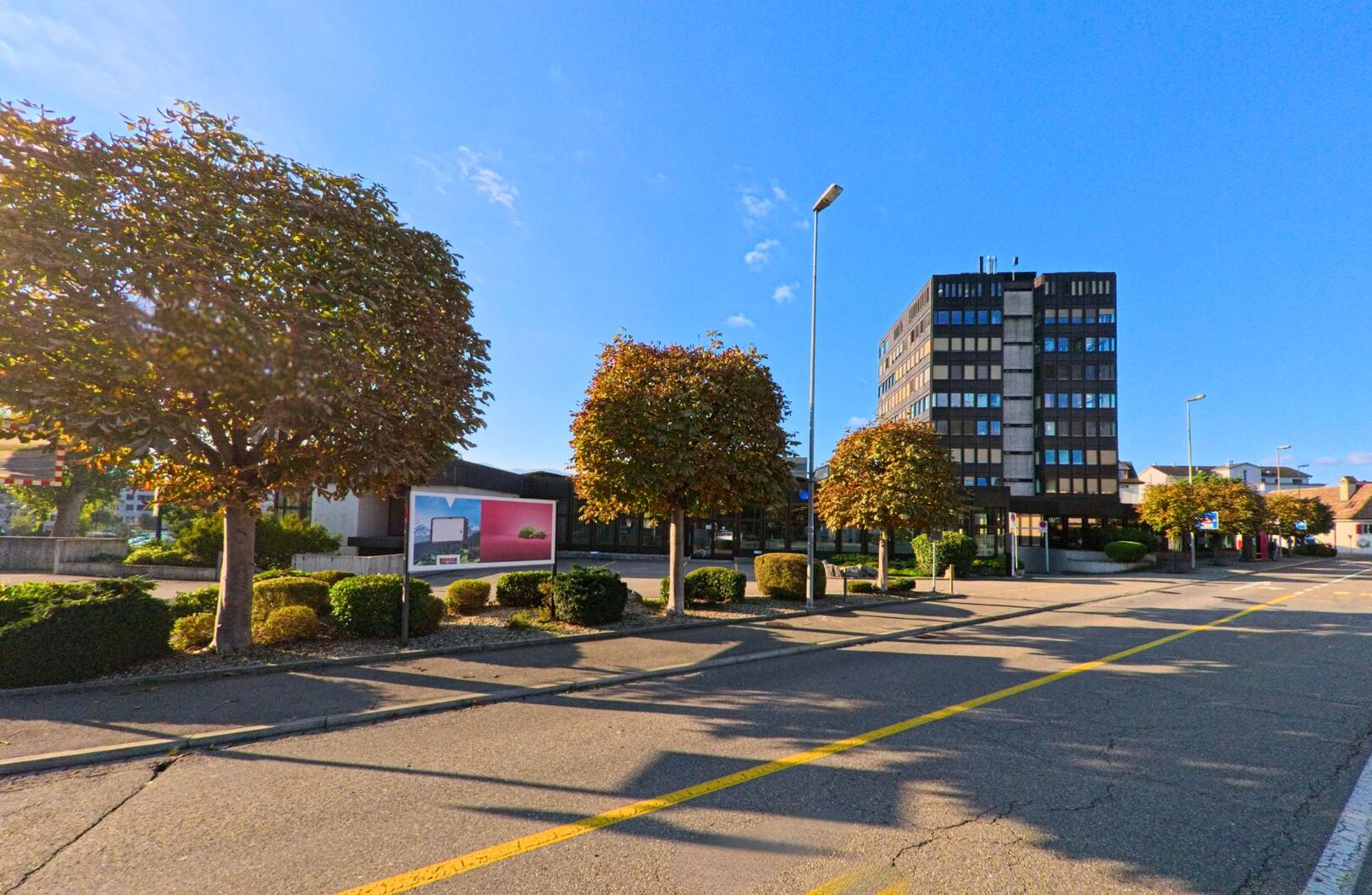 Vor einem mehrgeschossigen dunklen Gebaeude in Rapperswil-Jona (8640 im Kanton St. Gallen) verlaufen eine breite Strasse und eine Reihe geschnittener Baume im Sonnenlicht.