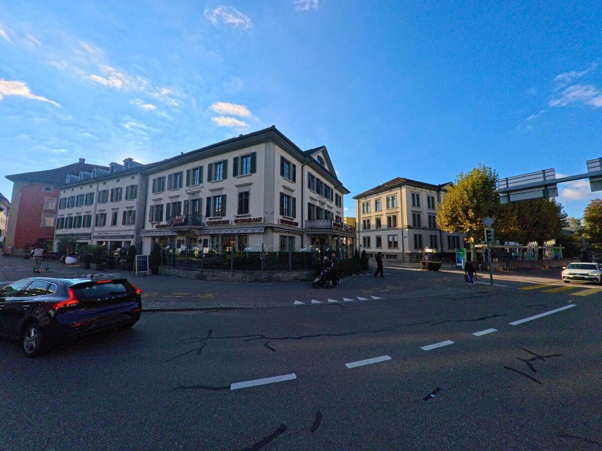 In Rapperswil-Jona (8640 im Kanton St. Gallen) stehen mehrere traditionelle Gebaeude an einer belebten Kreuzung, waehrend Passanten und Fahrzeuge den Strassenraum nutzen.