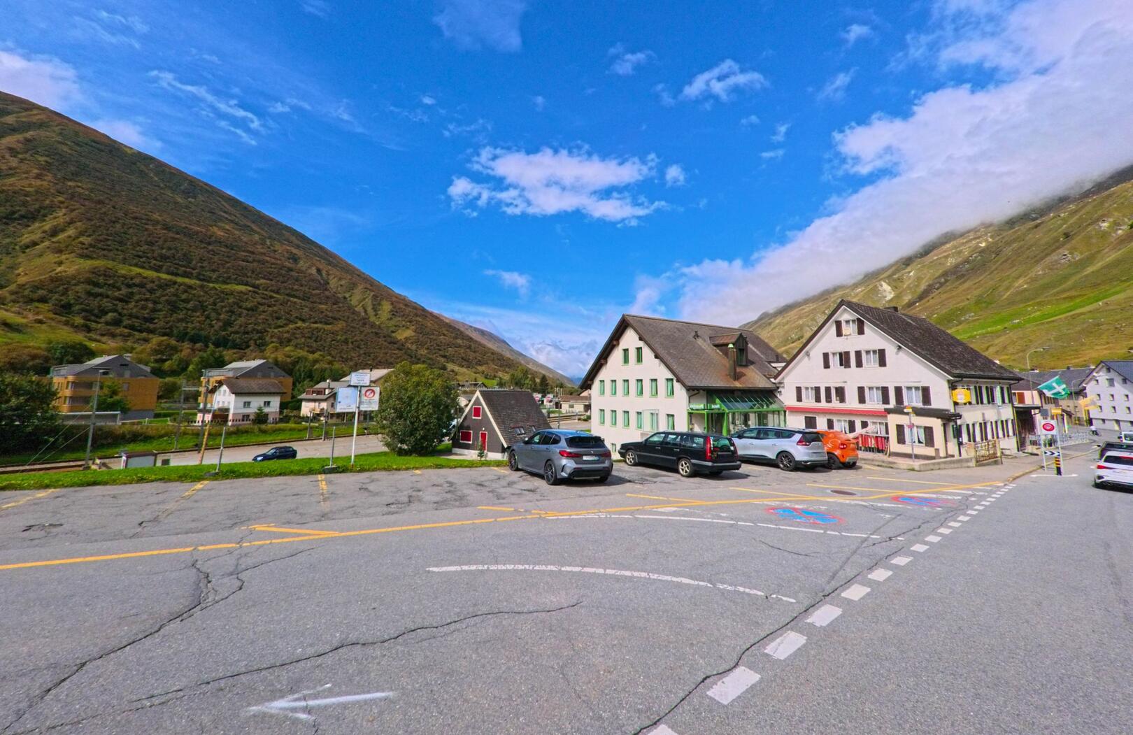 Vor den Berghängen in Realp (6491, Kanton Uri) stehen mehrere traditionelle Häuser mit Parkplätzen im Vordergrund unter blauem Himmel.