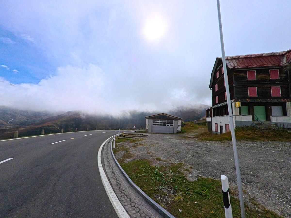 Am Rand von Realp (6491, Kanton Uri) führt eine Passstrasse an einem dunklen Gebäude mit roten Fensterläden vorbei, während Nebel über den Bergen liegt.