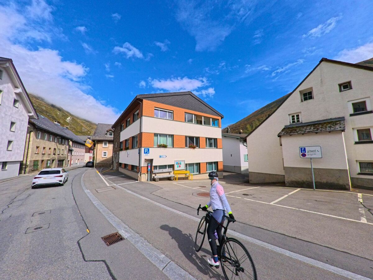 Eine Radfahrerin fährt durch eine schmale Strasse in Realp (6491, Kanton Uri), umgeben von Wohnhäusern mit Berglandschaft im Hintergrund.