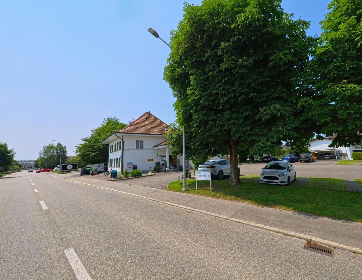 Ein Gebäude in Recherswil (4565, Kanton Solothurn) steht an einer breiten Straße, umgeben von grünen Bäumen und einem Parkplatz mit mehreren geparkten Fahrzeugen.