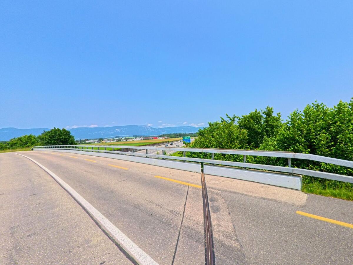 In Recherswil (4565, Kanton Solothurn) führt eine kurvige Straße entlang von grünen Büschen mit einem weiten Blick auf das Umland und die Alpen im Hintergrund.