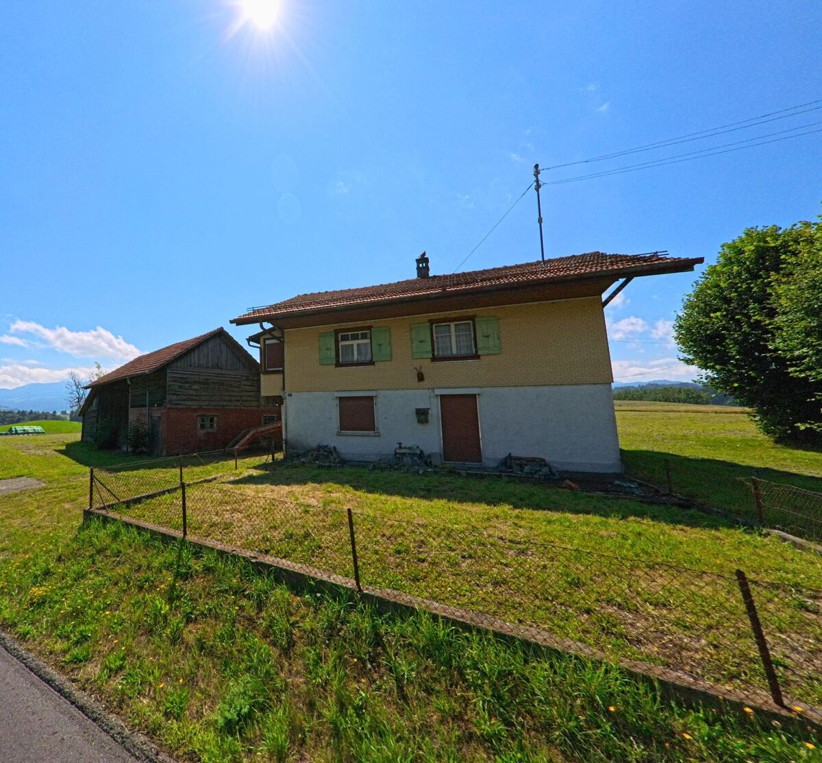 Am Rand einer Wiese in Rechthalten (1718, Kanton Freiburg) steht ein zweistoeckiges Wohnhaus mit gruener Fensterlaeden neben einer alten Scheune.