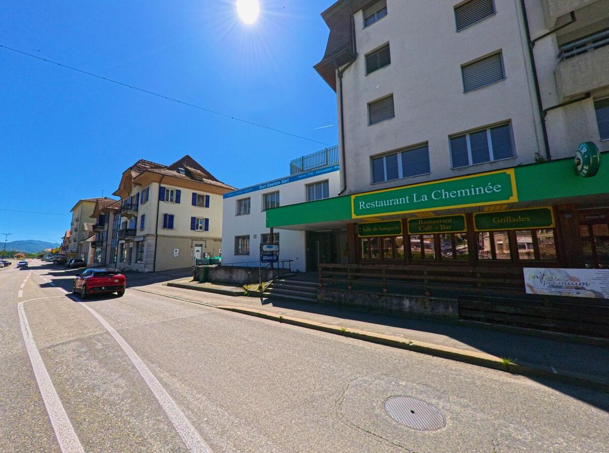 In Reconvilier (2732, Kanton Bern) befindet sich das Restaurant La Cheminée an einer sonnigen Hauptstrasse, flankiert von weiteren Wohn- und Geschäftshäusern.