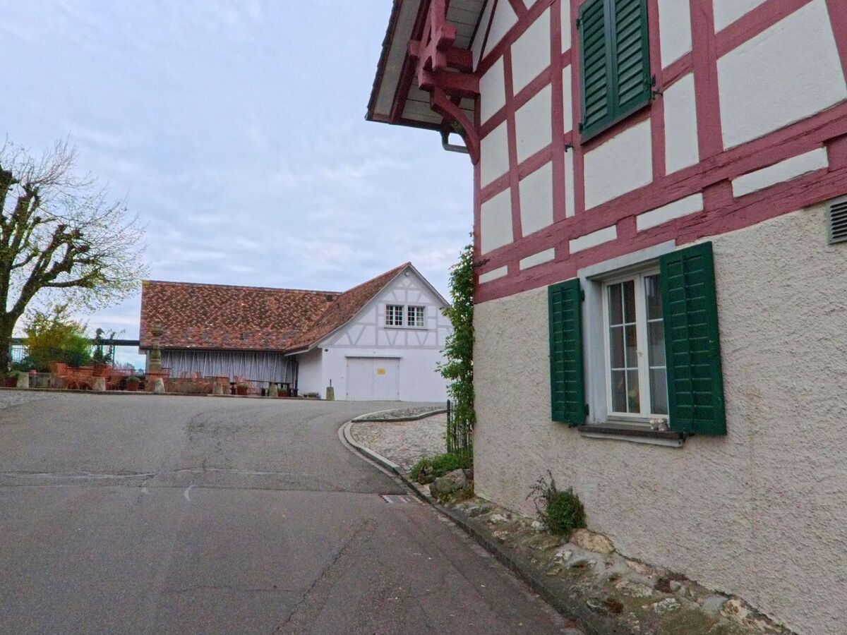 Zwei Fachwerkhäuser mit grün gestrichenen Fensterläden prägen diese ruhige Strassenszene in Regensberg, PLZ 8158 im Kanton Zürich, nahe einer kleinen Platzfläche mit Sitzgelegenheiten.