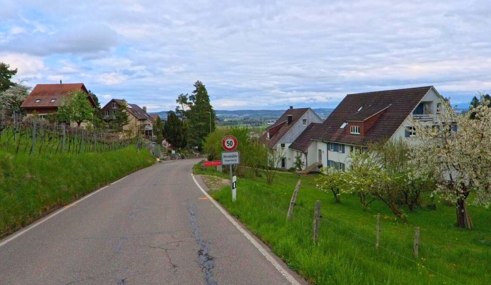 Auf dem Bild ist die Ortseinfahrt zur Siedlung Hirsmühle in Regensberg, PLZ 8158 im Kanton Zürich zu sehen, mit Wohnhäusern links und rechts der Landstrasse und Blick auf das Zürcher Unterland.