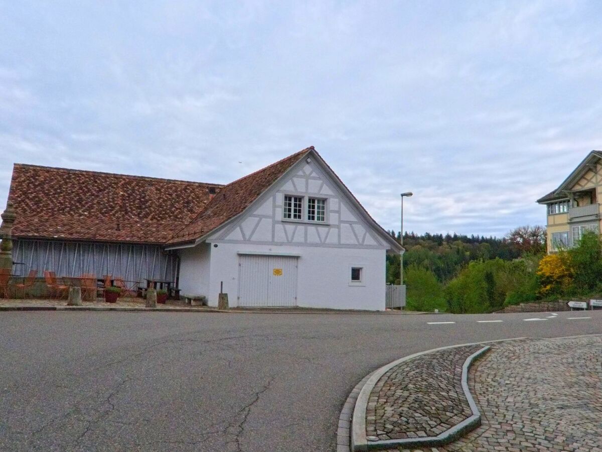 Ein weiss gestrichenes Fachwerkhaus mit Ziegeldach steht an einer gepflasterten Strassenkurve in Regensberg, PLZ 8158 im Kanton Zürich, mit Blick auf bewaldete Hügel im Hintergrund.