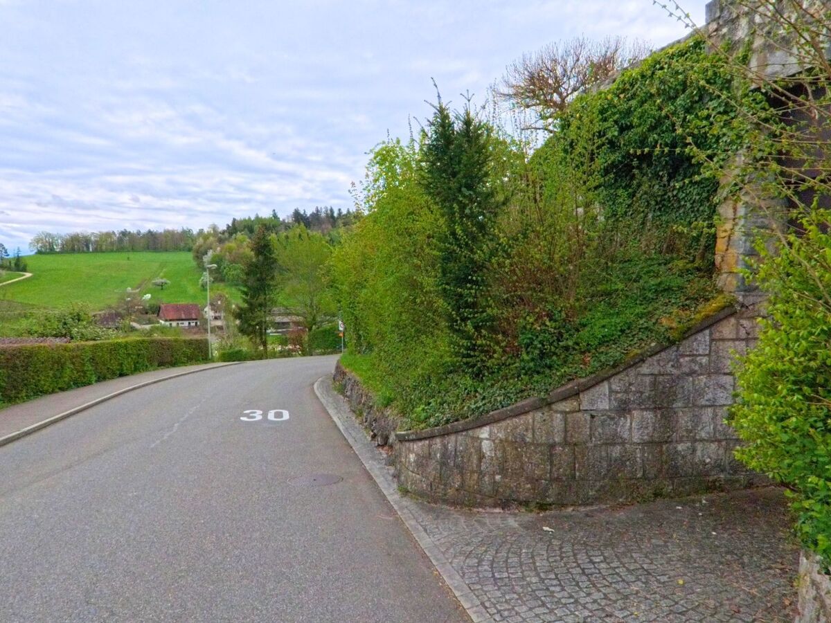 In Regensberg, PLZ 8158 im Kanton Zürich, verläuft eine Tempo-30-Zone entlang einer Kurve mit begrünten Mauern und Ausblick auf das ländlich geprägte Umland mit verstreuten Höfen.