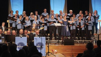 Der Regi-Chor Muri (5630, Kanton Aargau) tritt gemeinsam mit einer Big Band auf einer hell ausgeleuchteten Bühne unter der Leitung einer Dirigentin auf.