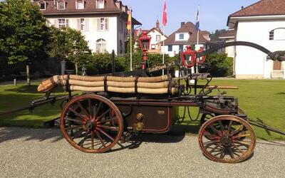 Eine historische Feuerwehrspritze der Regiowehr Suhrental Oldies steht in Schöftland (5040, Kanton Aargau) mit aufgerollten Schläuchen und roten Laternen vor einem öffentlichen Gebäude.