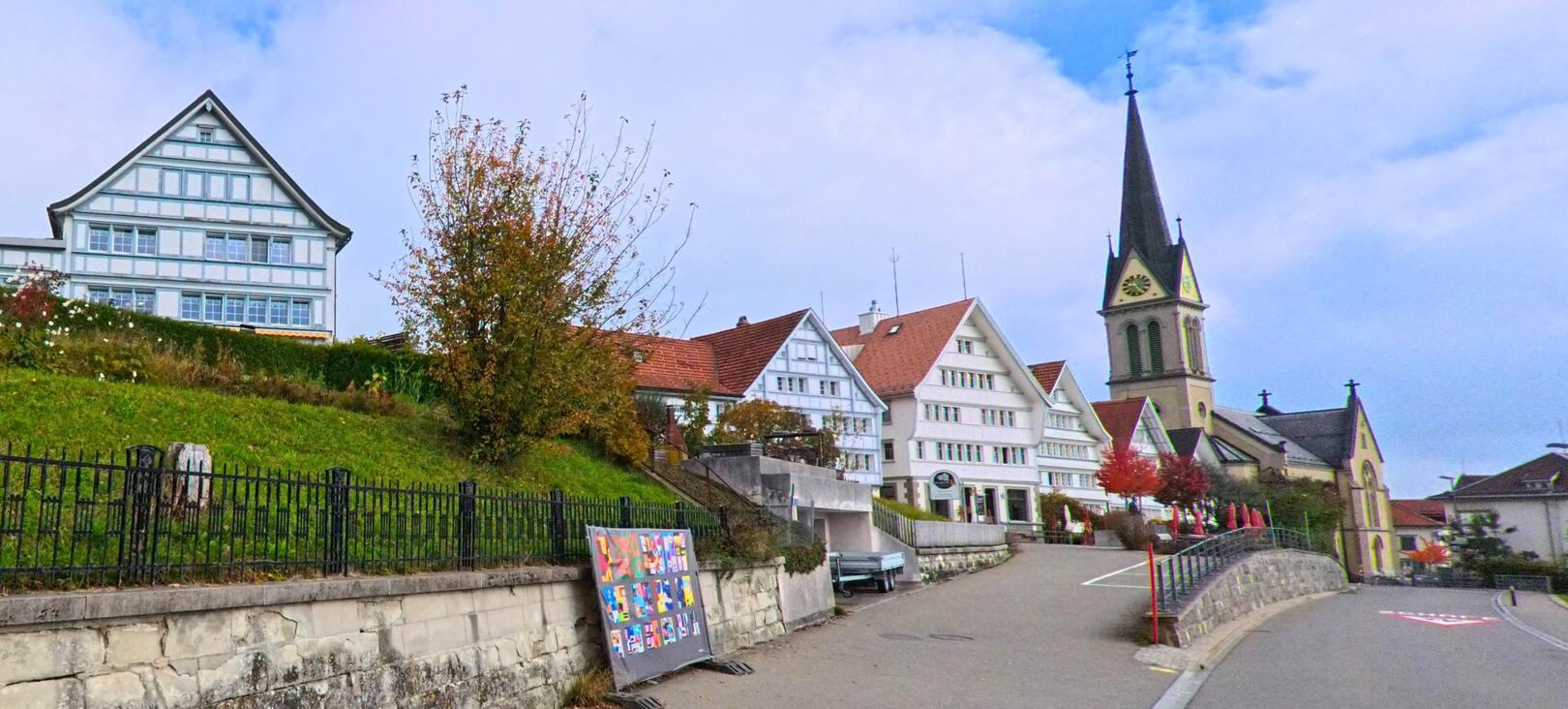 In [ORT] (PLZ [PLZ], Kanton [KANTON]) saeumen traditionelle Holzhäuser und die Kirche mit dem markanten Turm eine leicht ansteigende Strasse unter herbstlichem Himmel.