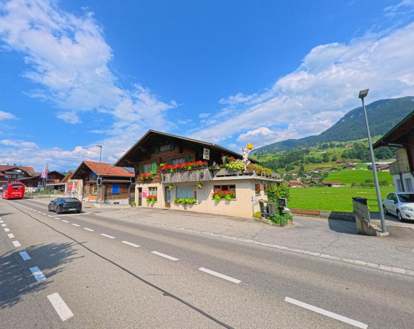 In Reichenbach im Kandertal (3713, Kanton Bern) säumen verzierte Häuser mit Blumenschmuck die Hauptstrasse, während Autos und ein roter Bus durch das sonnige Tal rollen.