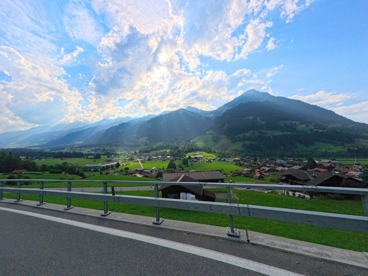 Von einer erhöhten Strasse in Reichenbach im Kandertal (3722, Kanton Bern) öffnet sich der Blick über das Tal mit verstreuten Häusern und dramatischer Wolkenkulisse über den Bergen.