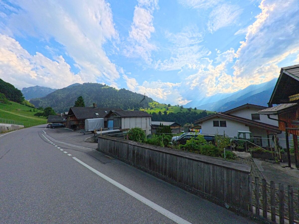In Reichenbach im Kandertal (3722, Kanton Bern) schlängelt sich eine Strasse an Wohnhäusern und Schuppen vorbei durch eine grüne Hügellandschaft mit eindrucksvoller Wolkenstimmung.